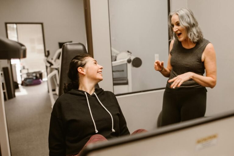 Two women in a gym setting sharing a joyful moment, promoting fitness and wellbeing.